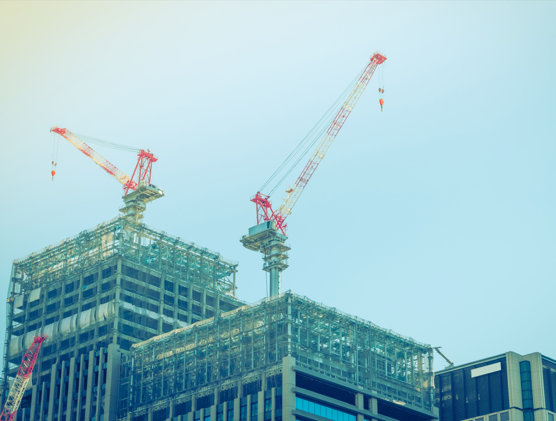 Two cranes tower over unfinished skyscrapers against a pale blue sky.