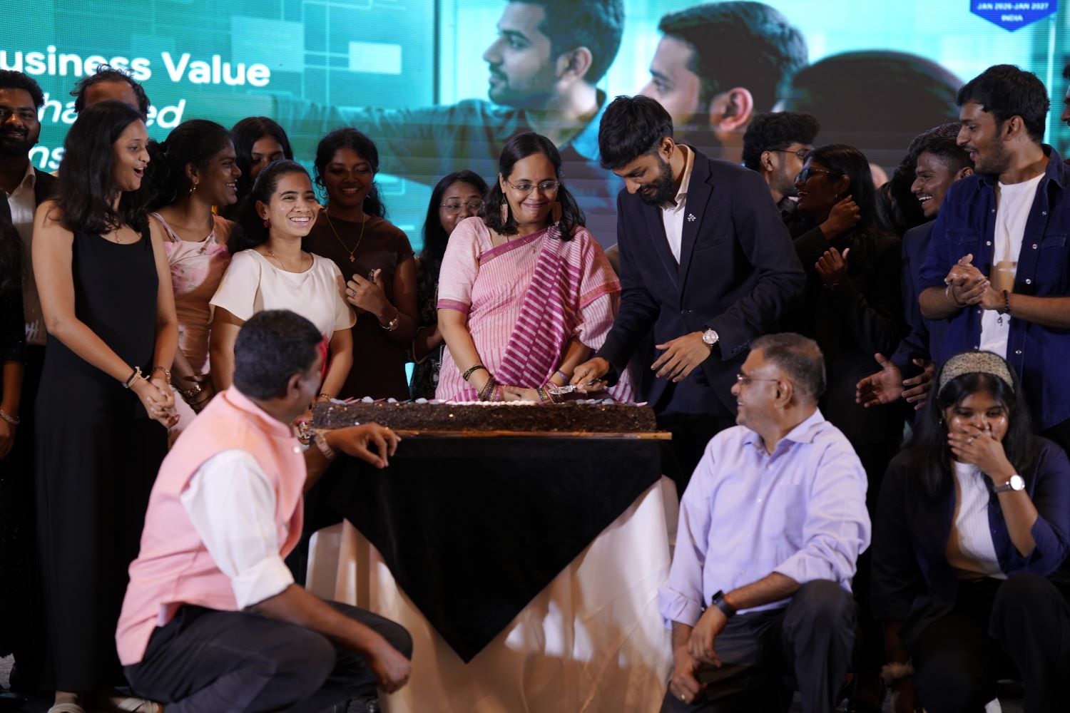 Group of people celebrating with a cake, with a man in a suit cutting it.