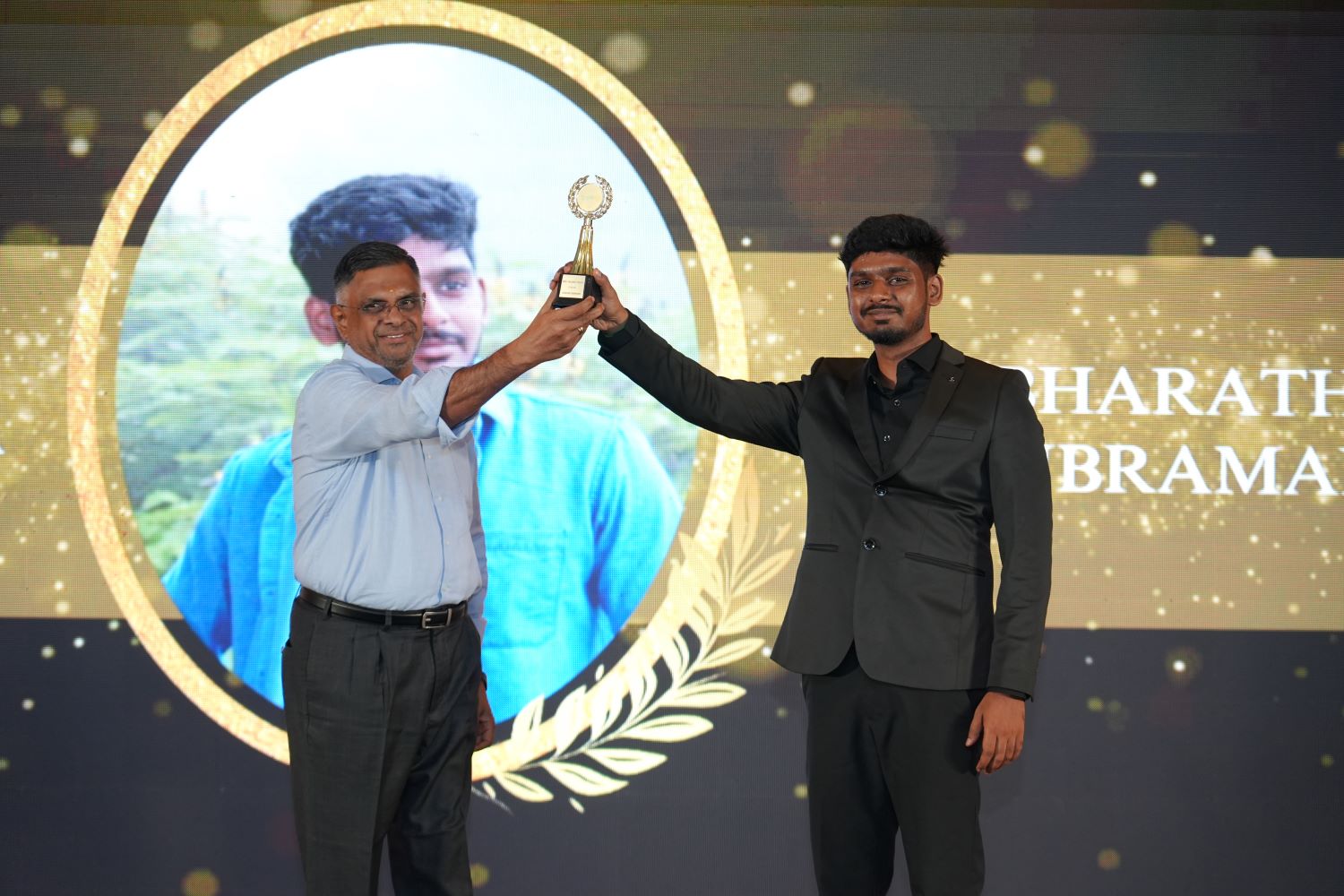Two men on stage, one presenting a trophy to the other. Text on screen reads HARATH and BRAMA.