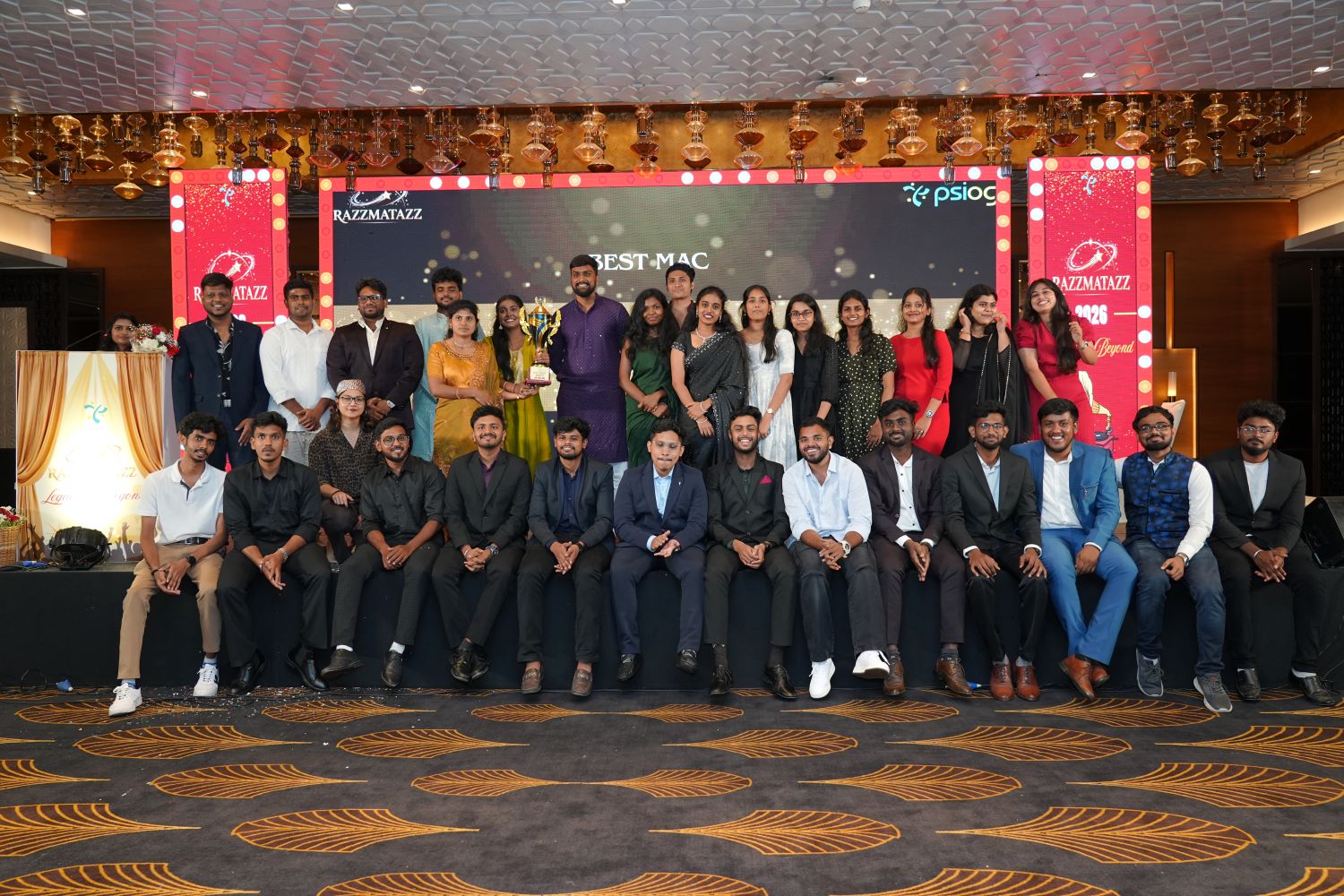 Group photo of award recipients posing for a picture on stage with a large screen displaying BEST MAC.